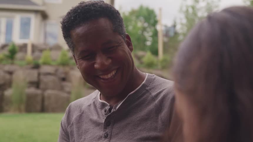 A man smiles and laughs as he talks with his female companion outdoors near a house