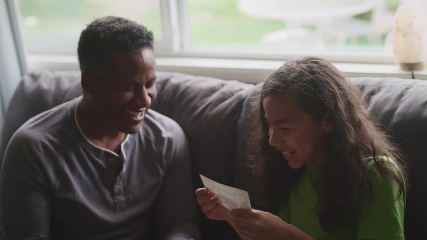 Father and daughter sitting in a couch in the living room laugh while reading a card