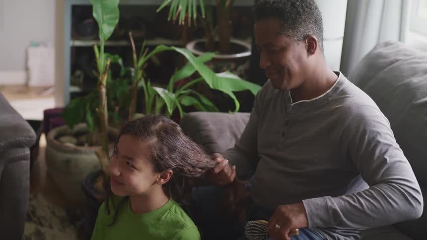 A man sitting on a couch brushing a girls hair with a brush