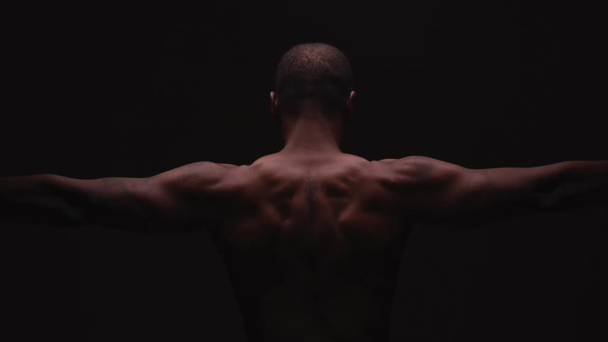 Back of a shirtless muscular man extending his arms inside a dark room
