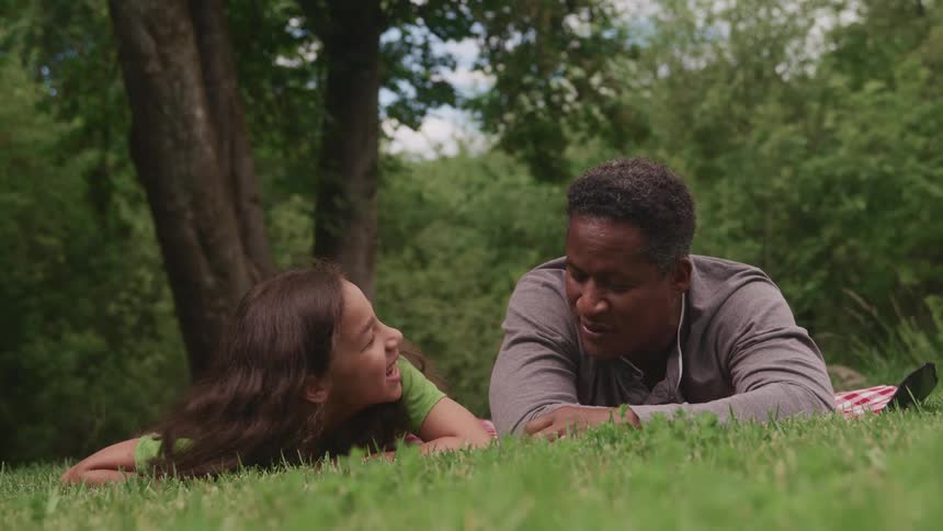 Man and girl lie in the green grass and talk to each other with trees in the background