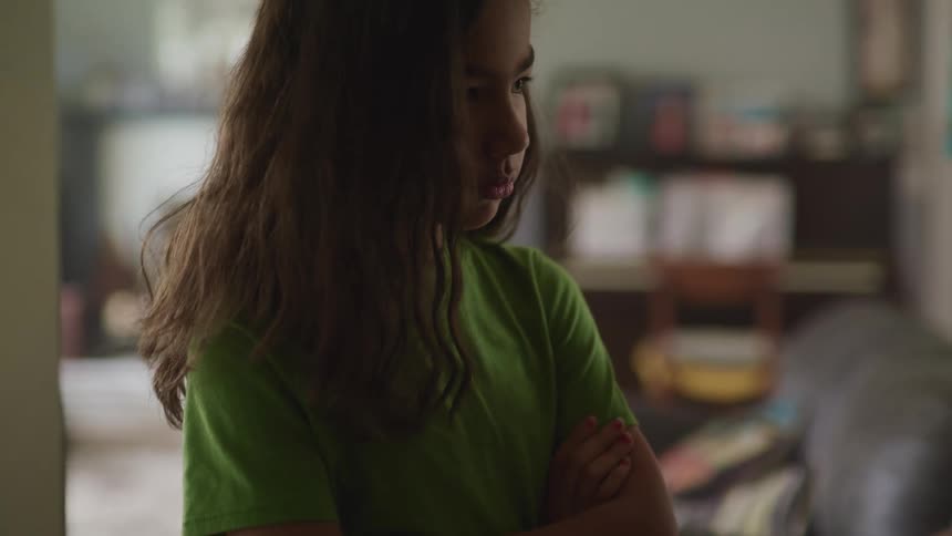 A girl standing with her arms crossed in the living room of a house raises her head to look at a man standing next to her