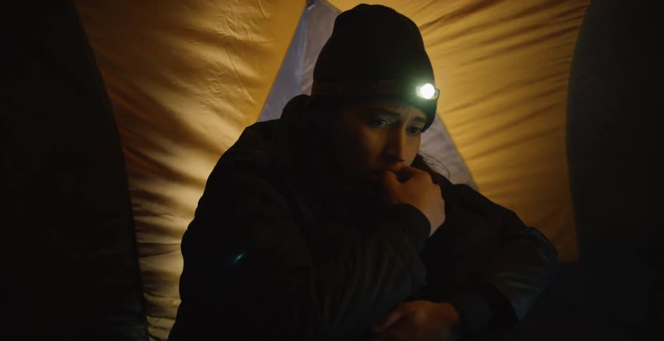 A person sits in a tent wearing a head lamp hat and coat while the wind blows and shakes the tent