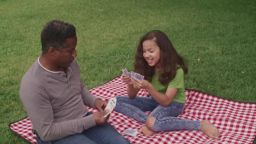 A man and a girl sitting on a blanket on grass playing cards