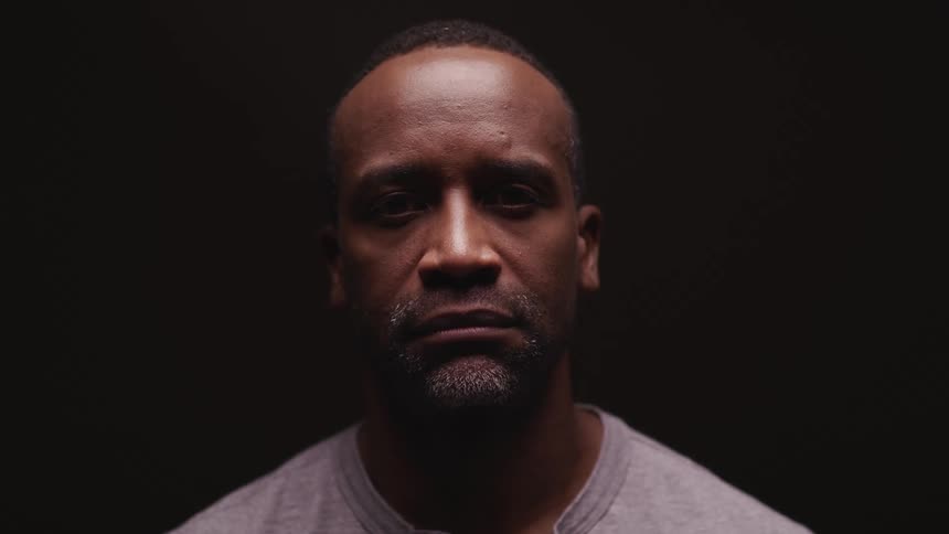 Man with a goatee raises his head to look at the camera on a dark background