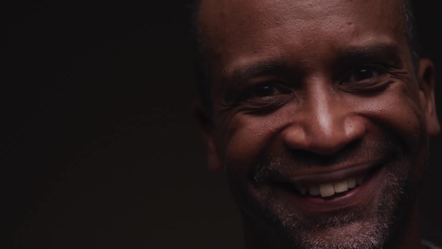 A black man looks up smiling and laughing against a black background