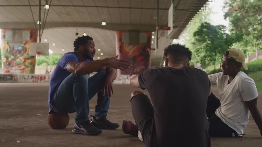 Three men sitting on a basketball court and talking with trees the sky and clouds in the background
