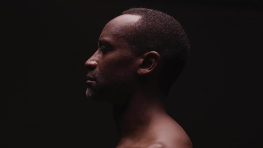 A man in a black room looking to the left lowers his head and shuts his eyes