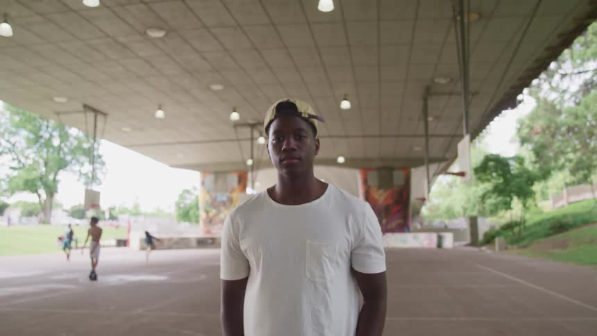 A young man stares into the camera while standing on an outdoor basketball court under a bridge during the day