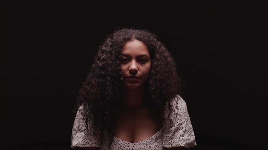 A young woman sitting in a dark room raises her head to look into the camera