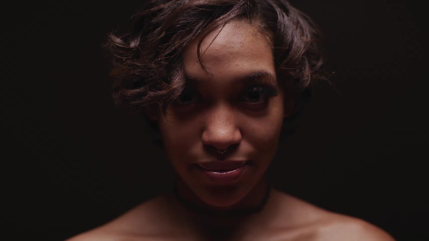 A woman stands in a dark room with her shoulders exposed and smiles as she looks up
