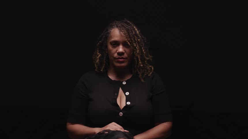 A woman in a black room lifts her head and stares at the camera