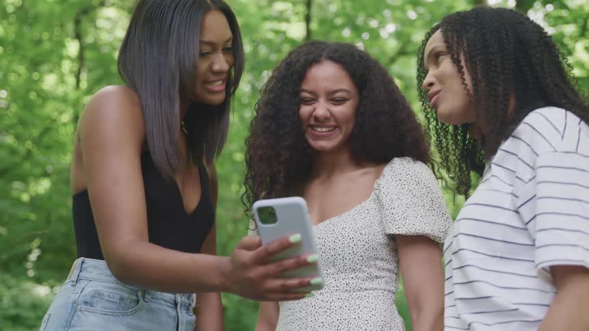 Three women looking at a smart phone with trees the sky and clouds in the background