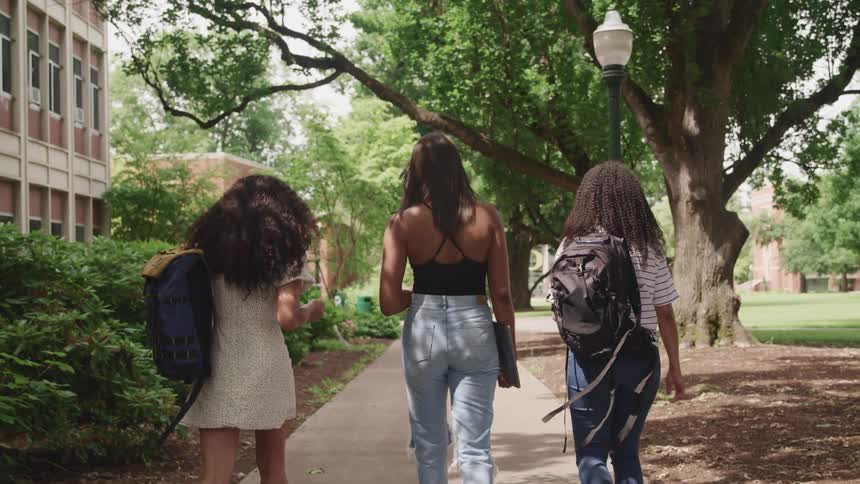 Three women walk side by side on a campus path on a sunny day