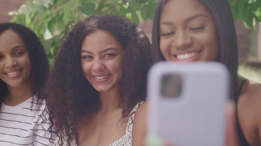 Three women taking a picture with a smart phone with leaves grass and a building in the background
