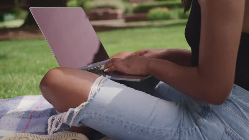 Woman sits cross legged on blanket on lawn typing on laptop set on her lap