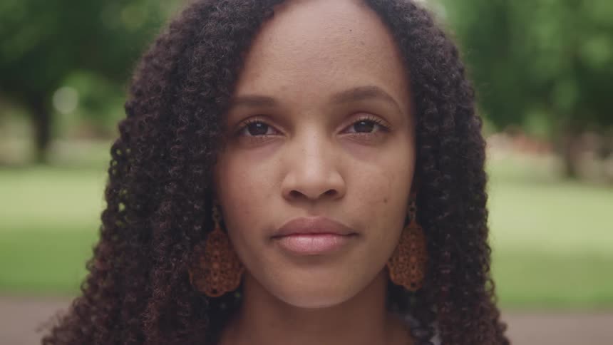 A woman with long curls and big earrings looks straight ahead with trees and grass behind