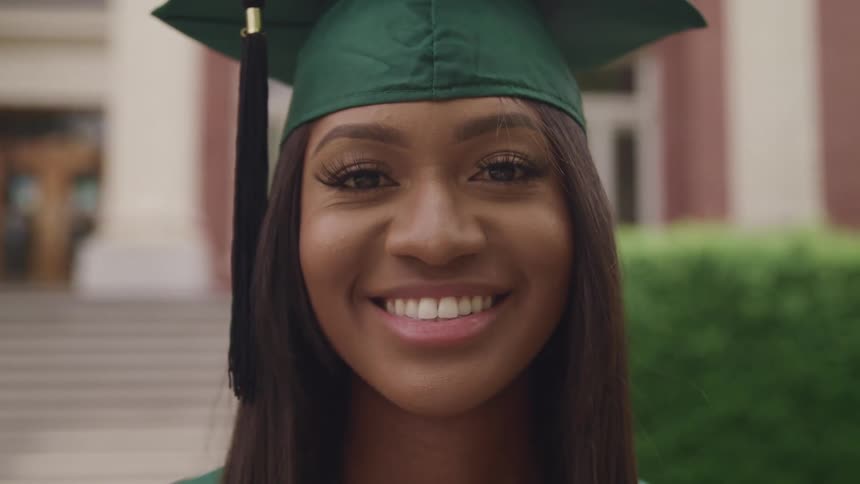A woman with a graduation cap and gown smiling with a building and a hedge in the background