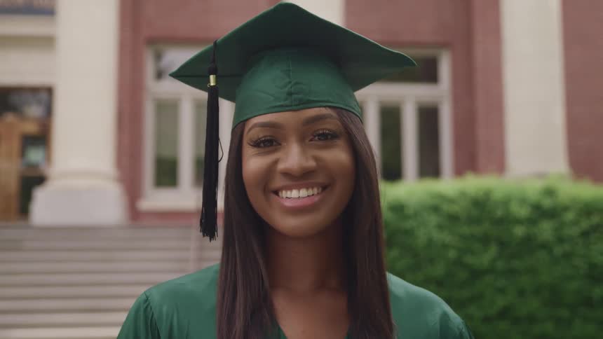 Woman wearing green graduation gown stands near steps of building smiling widely