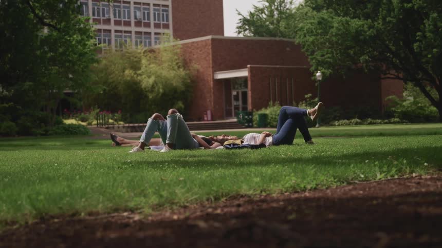 Women laying on grass with a building trees the sky and clouds in the background
