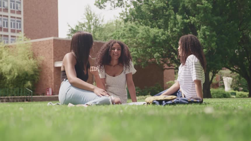 Three female friends smiling and laughing while sitting on a grassy lawn