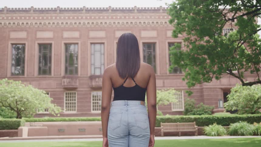 A woman with jeans standing with a building grass hedges and trees in the background