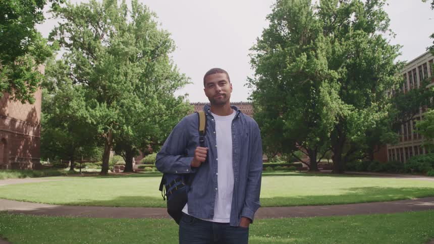 A man stands holding his backpack on a green campus with brick buildings behind