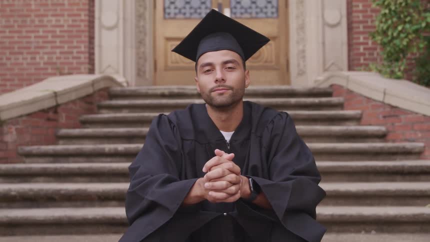 A man in academic regalia turns his head to look into the camera while sitting on a stone staircase outside of a building during the day