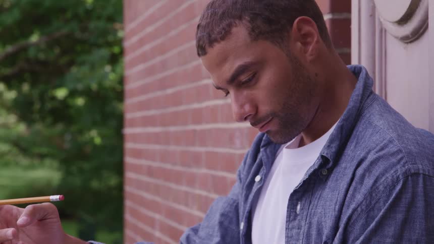 Man fiddles a pencil leaning against a wall next to a building