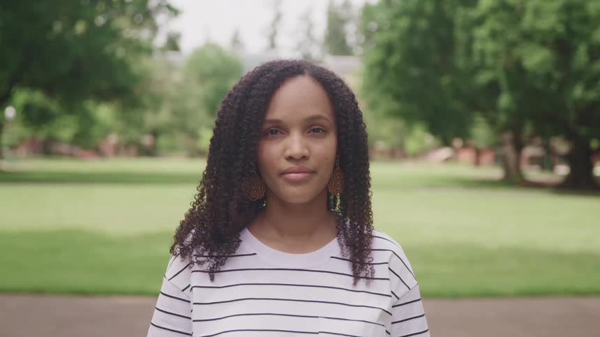 A woman in a striped shirt stands in front of a sidewalk in a public park