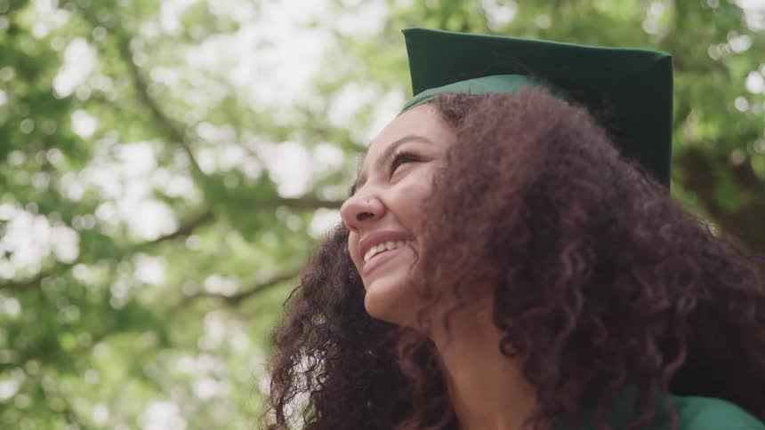A young woman wearing a green graduation robe smiling while standing outdoors under a tree at daytime