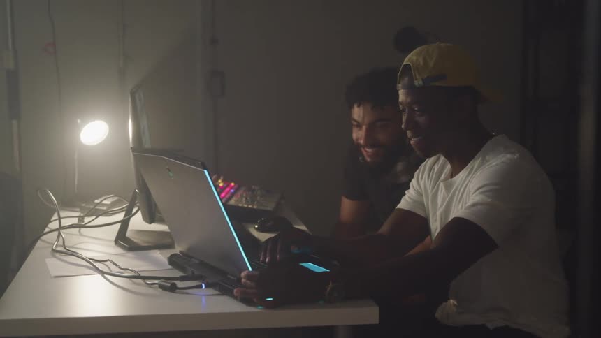 Two men smiling and laughing while using a laptop in a dimly lit room