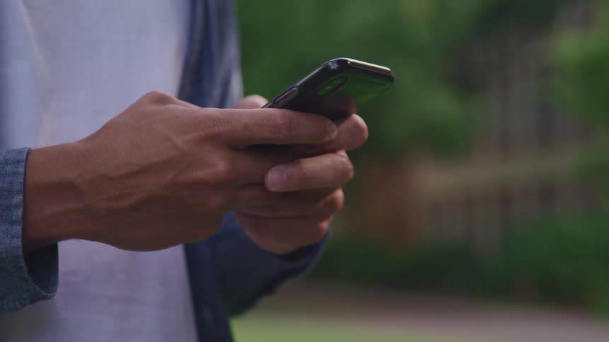 A man uses his thumbs to type on a smartphone while standing outdoors during the day