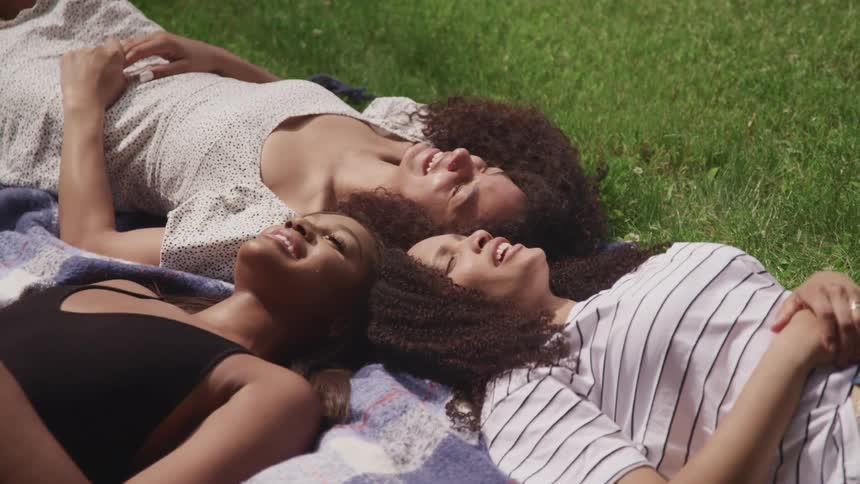 Three women smiling and talking while lying with their heads together on a grassy lawn