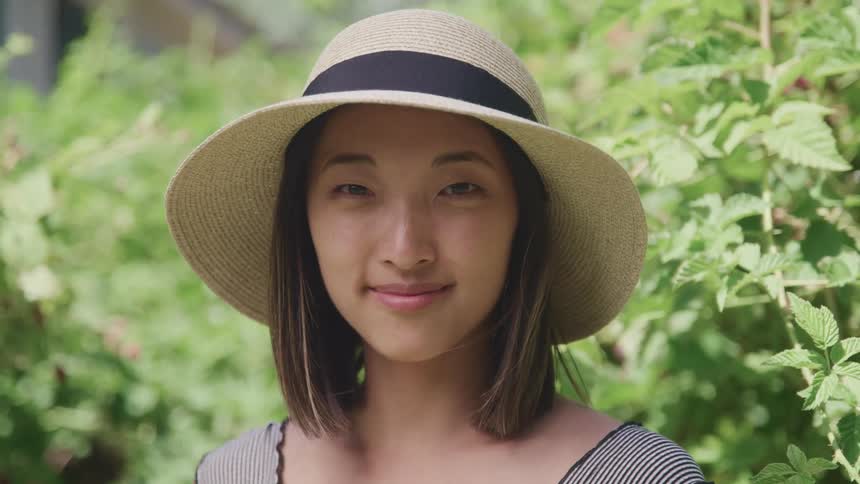 A woman with a hat standing and staring with branches and leaves in the background
