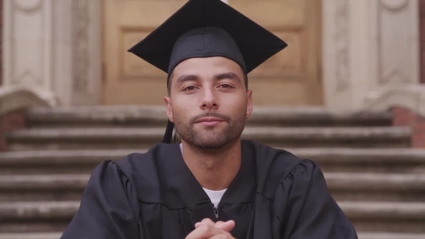 Young man wearing academic cap and gown sitting on the stairs of a building