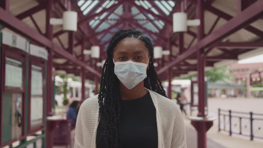 Woman with long braids wearing face mask stands near display board and sidewalk