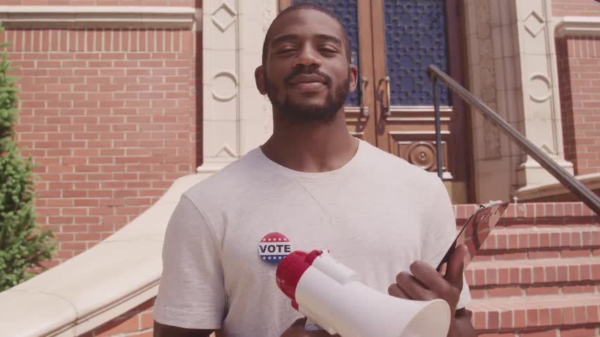 A black man stands proudly holding a megaphone and clipboard infront of a brick building with a vote button on his tee shirt