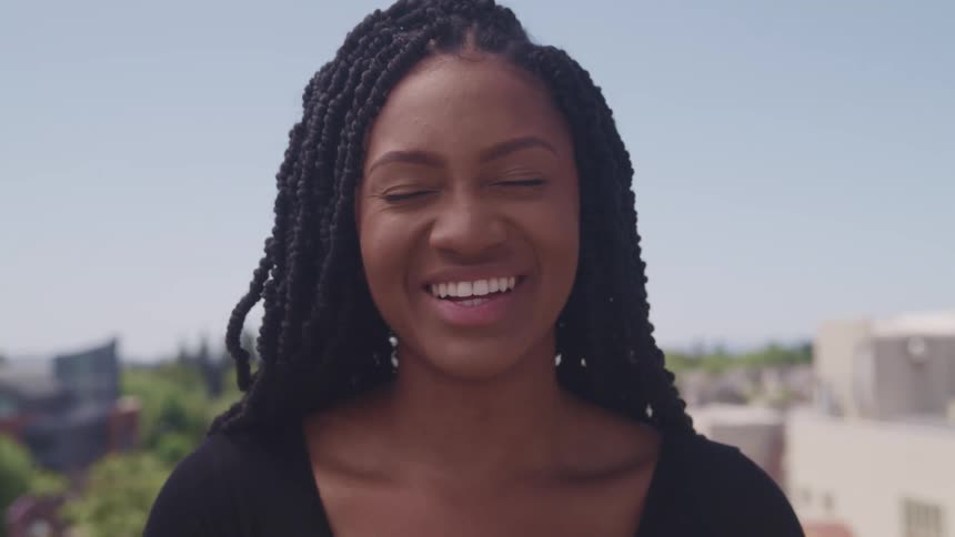 Woman with a black blouse and braids laughs and looks at the camera with houses in the background