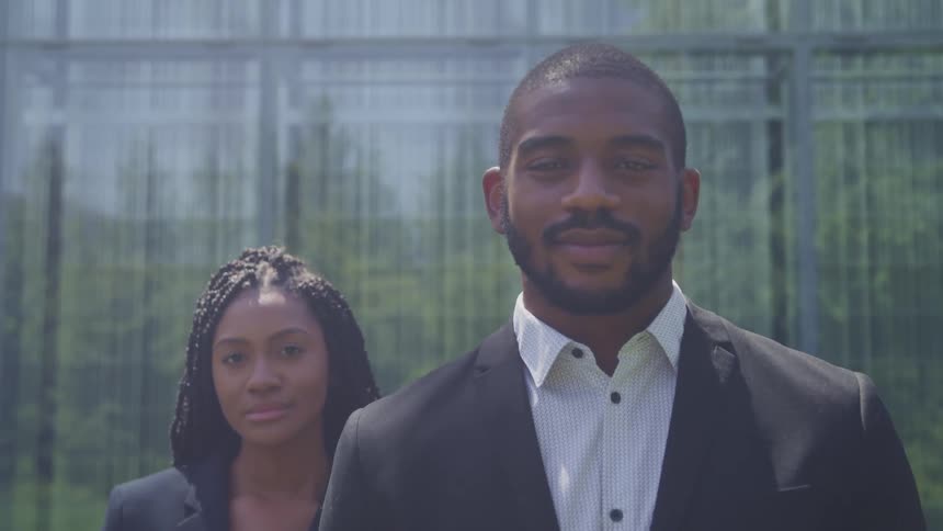 A woman stands behind a man while they both smile slightly and stare at the camera outdoors at daytime
