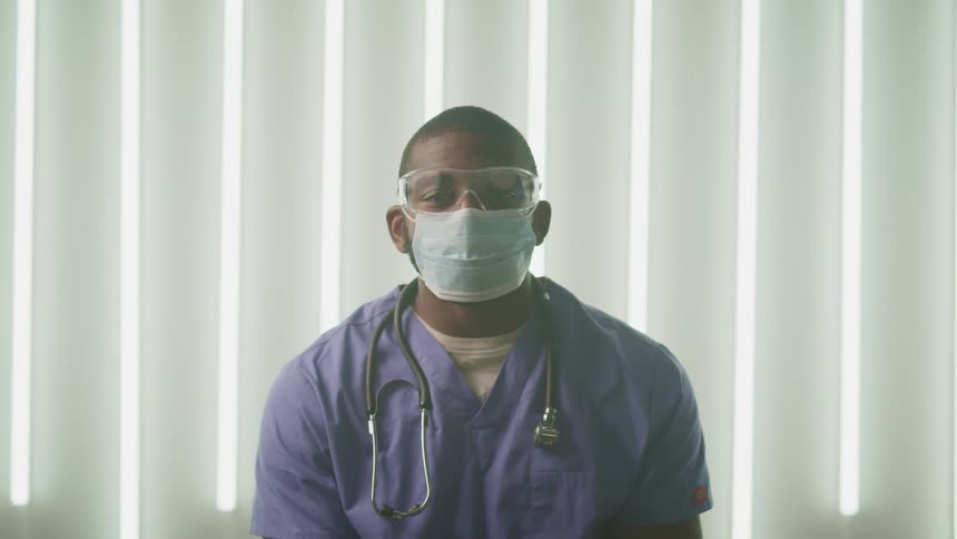 A man with goggles a mask a stethoscope and scrubs in a room with a window and blinds in the background