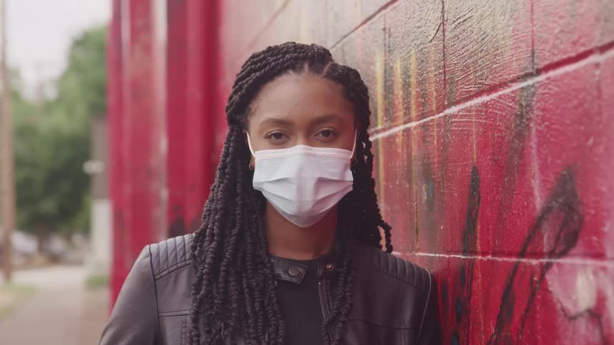A woman in a face mask poses next to a building painted red and marked with graffiti