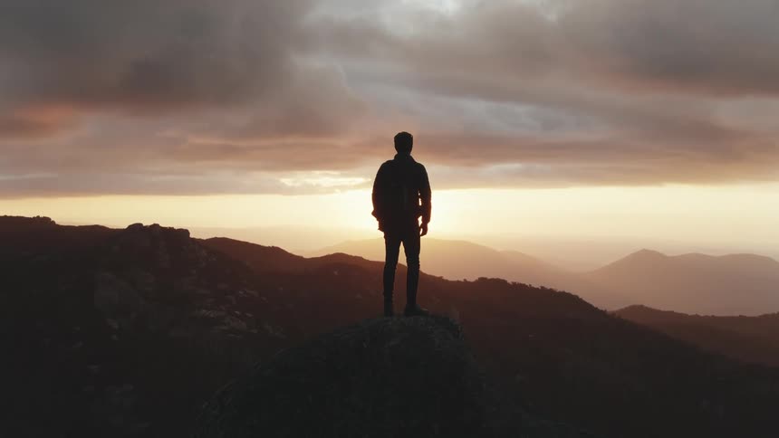 A man stands on a rock overlooking a mountain range at sunset