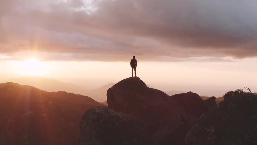 A man stands on top of a cliff as the sun sets behind him