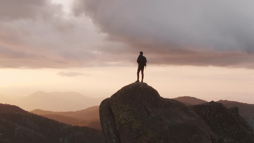 A man stands atop a large boulder on a ridge overlooking the mountains and hills on a cloudy day