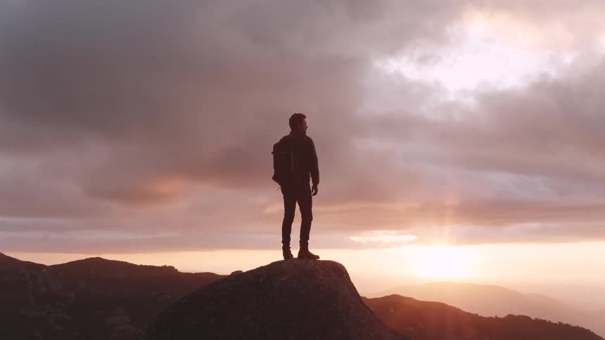 Man stands on big steep rock and looks around while the sun goes down