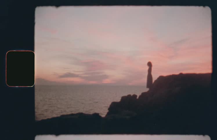 A woman in a dress stands on rocks on a coast and looks out to the ocean