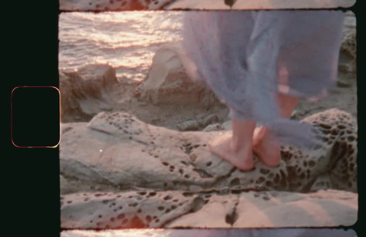 Vintage footage woman standing on rock overlooking coast as dress blows in the wind