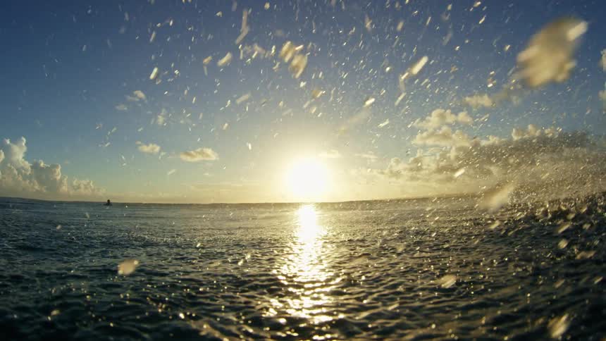 Water drops splash onto the ocean surface as sunlight shimmers and people swim and surf