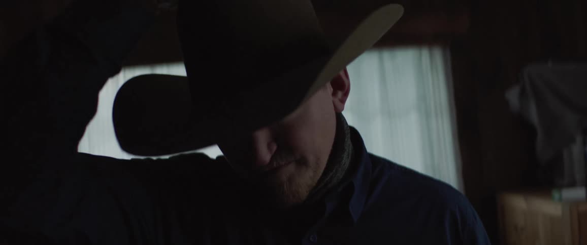 Man puts on cowboy hat standing in room with window at his back